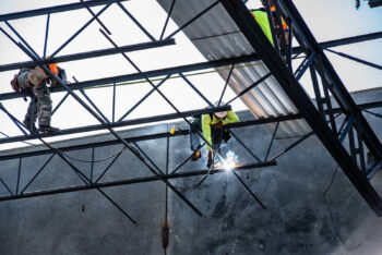 A man in a green shirt and a hardhat welds two bars of steel together suspended in the air